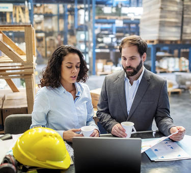 Two team members review information via laptop while standing in a warehouse